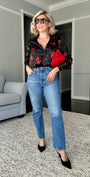 A woman modeling a black long-sleeve button-up shirt with red floral embroidery, paired with blue jeans, black pointed-toe heels, and sunglasses, standing in a modern living room setting.