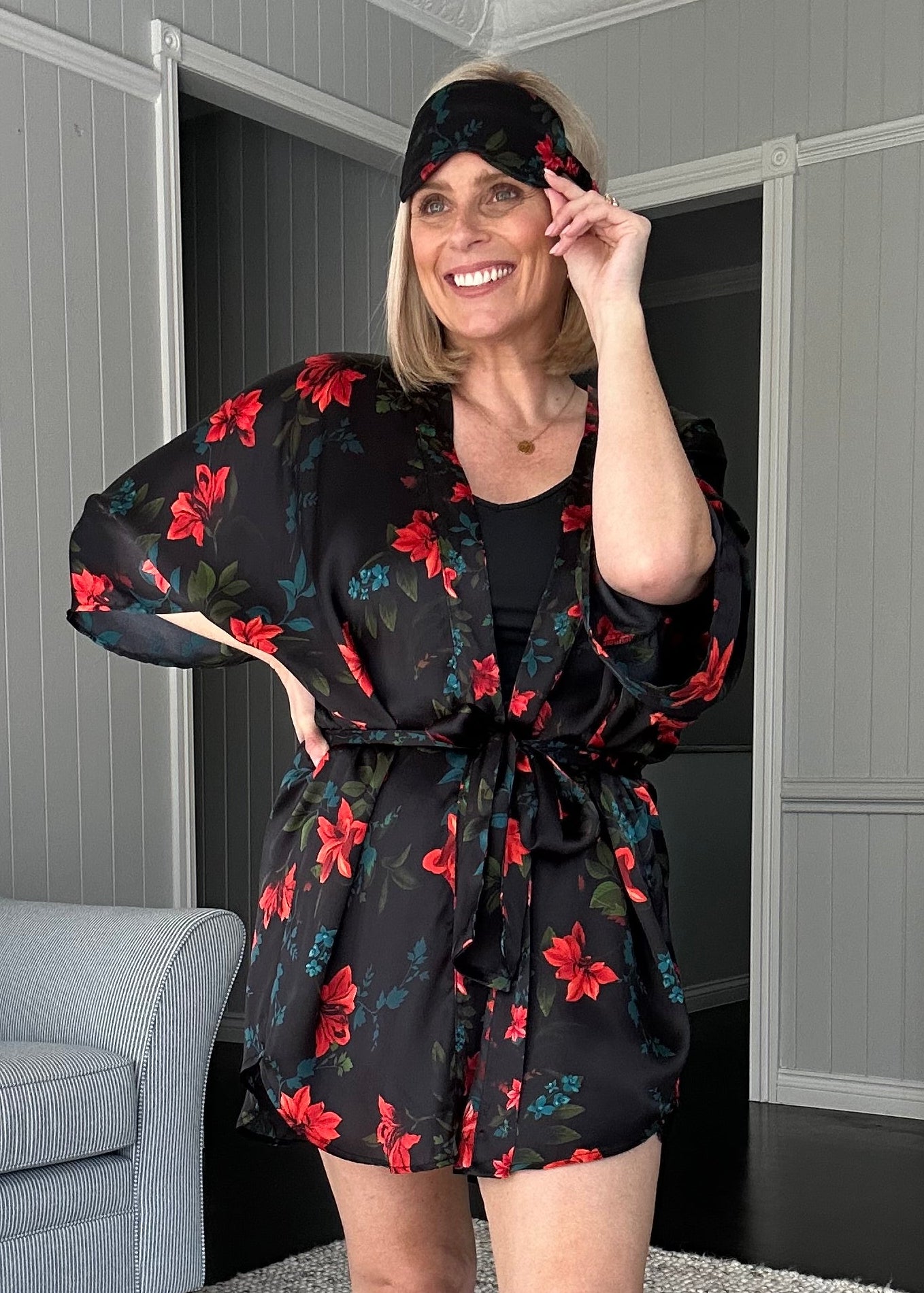 A woman wearing a black short-sleeve robe with a red floral pattern and tan slippers, posing in a modern living room setting.