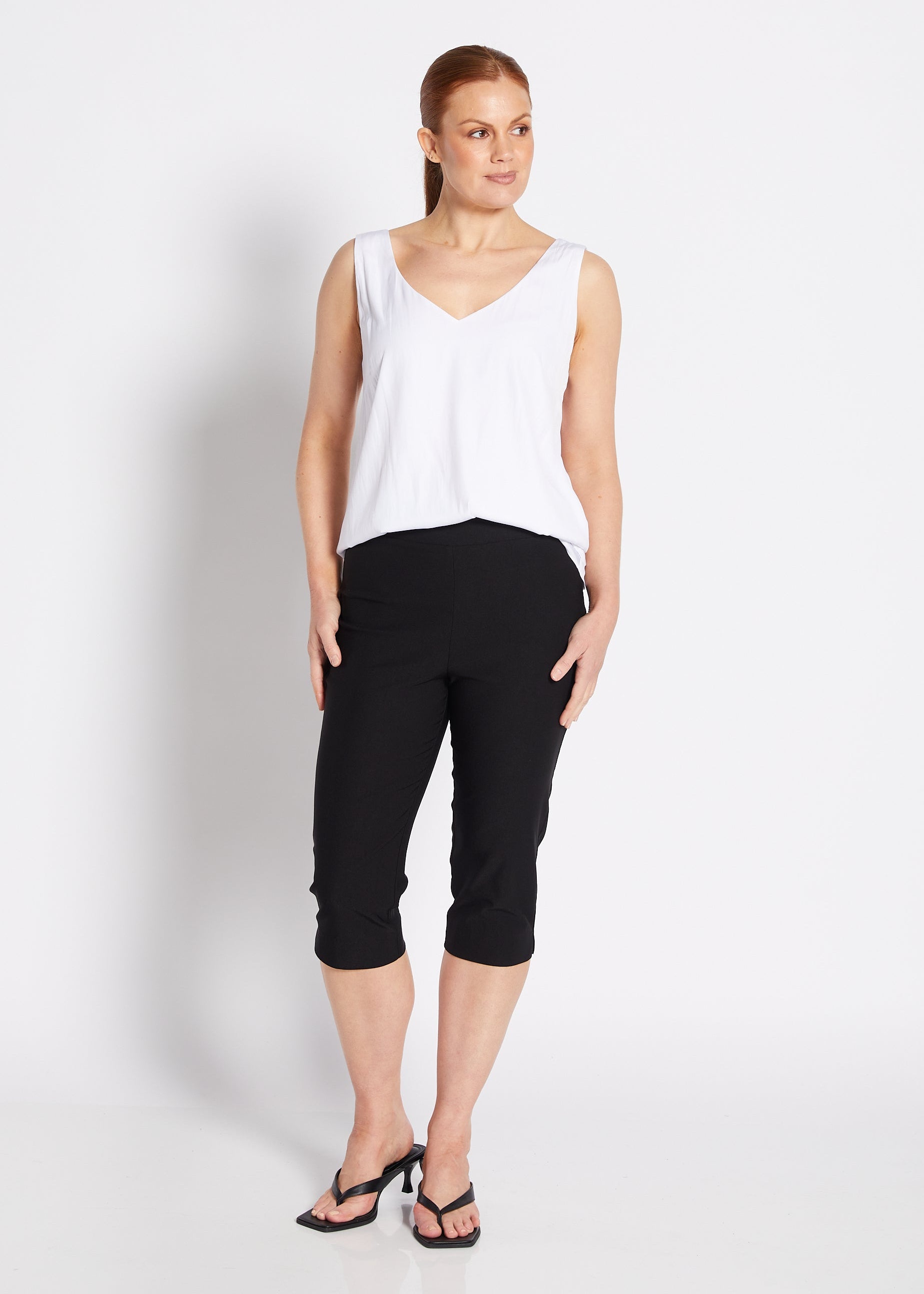 A woman modeling a white sleeveless V-neck top and black cropped pants against a plain white studio background.