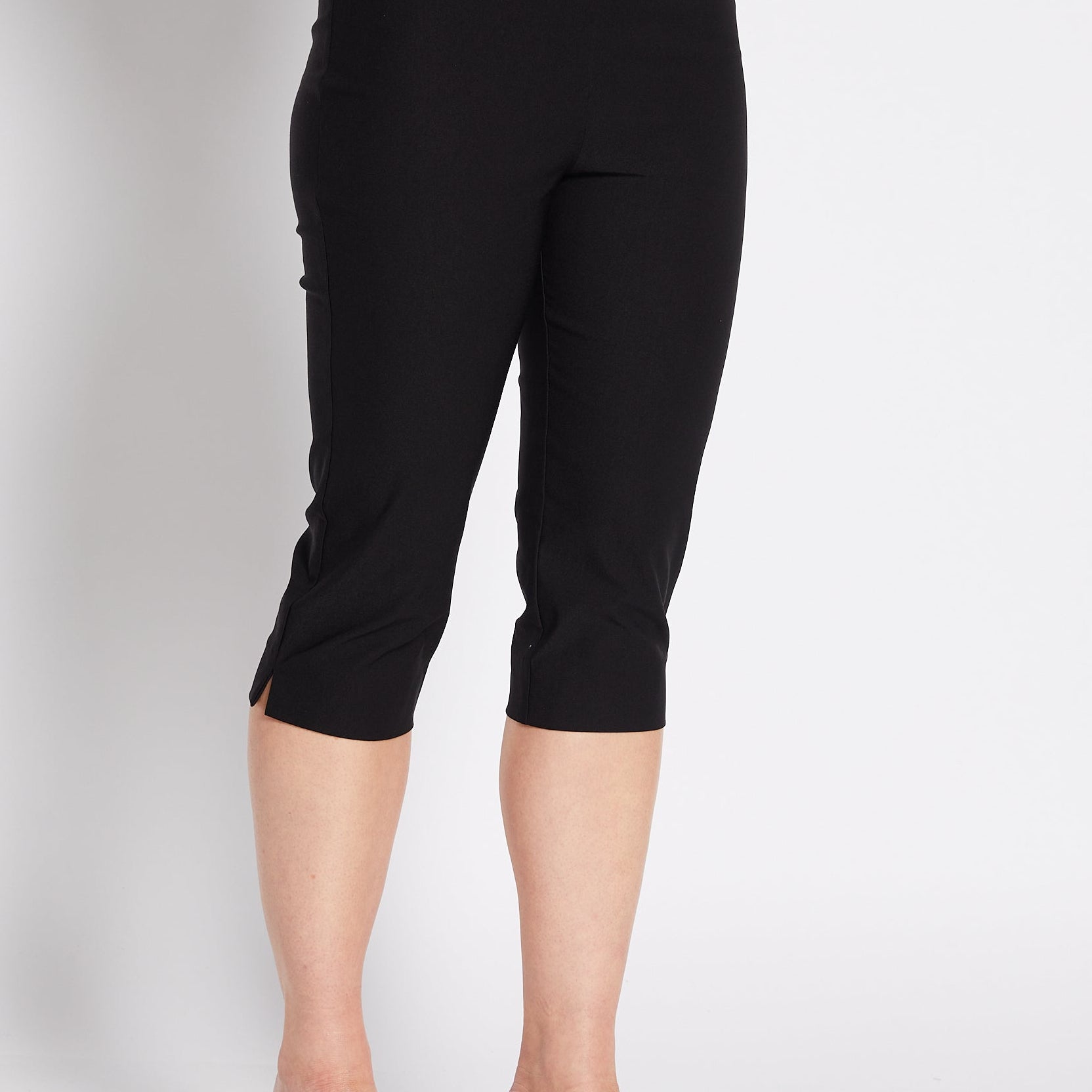 A woman modeling black capri pants with a white cropped tank top and black heeled sandals, posing against a light gray studio background.