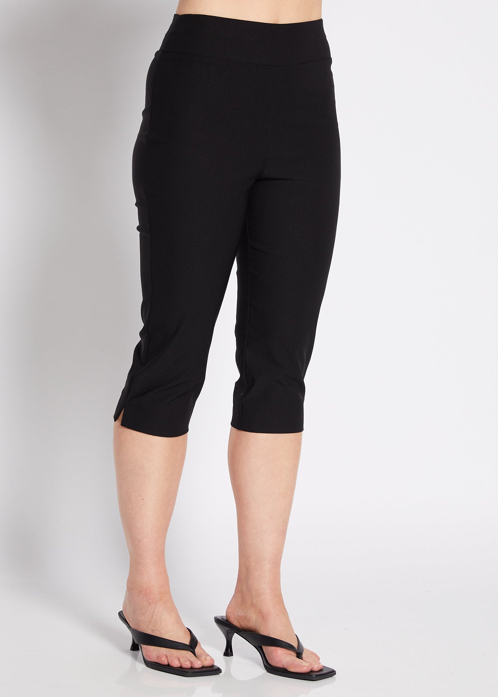 A woman modeling black capri pants with a white cropped tank top and black heeled sandals, posing against a light gray studio background.