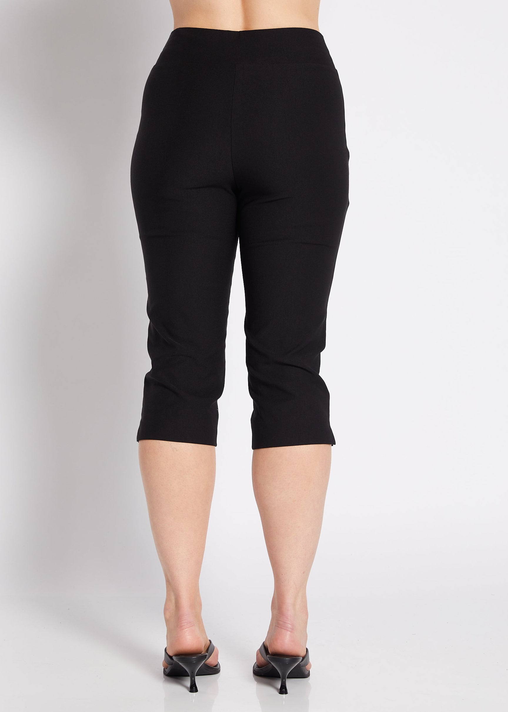 A woman shown from the back wearing black capri pants, a white tank top, and black pointed heels against a plain light gray studio background.