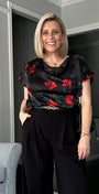 A woman modeling a black wide-leg pants set with a red floral print blouse in a modern living room setting.