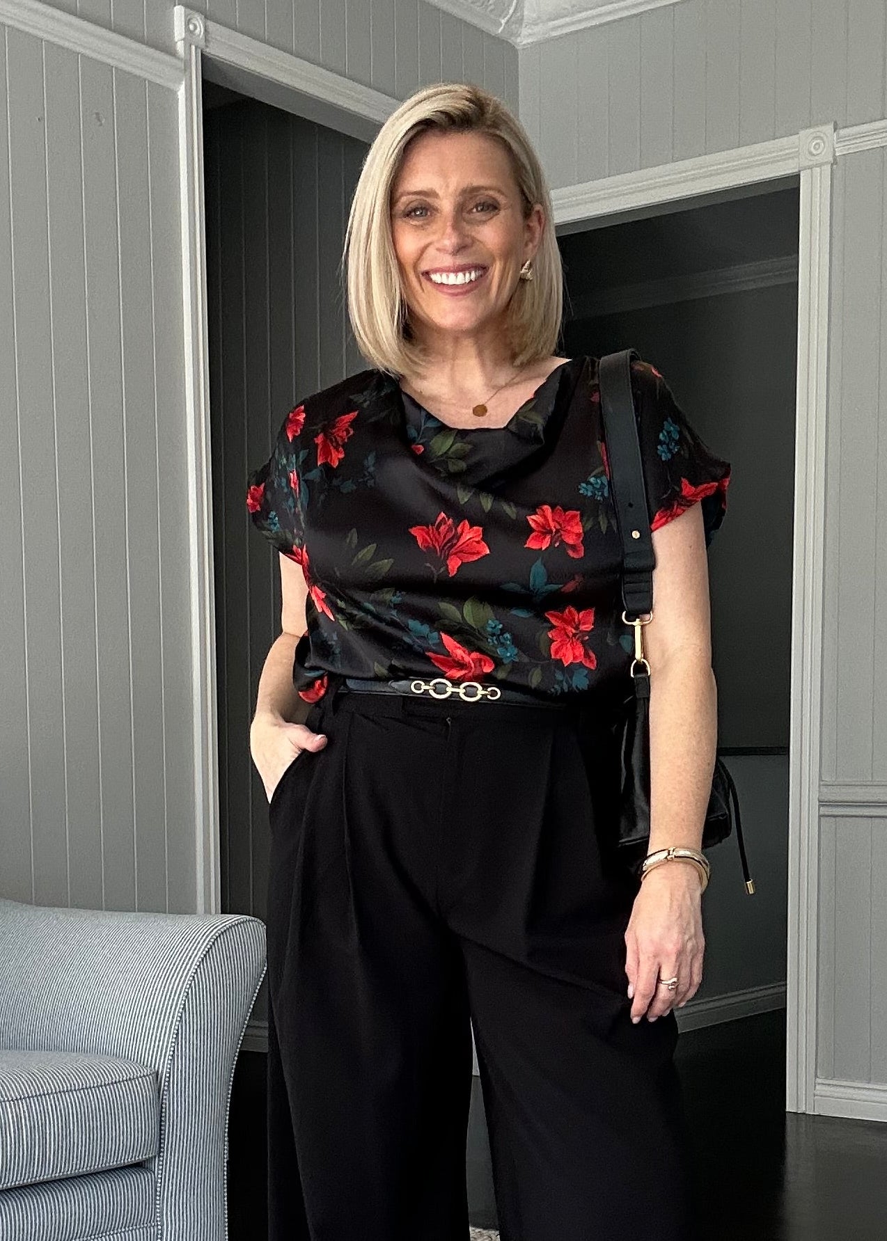 A woman modeling a black wide-leg pants set with a red floral print blouse in a modern living room setting.