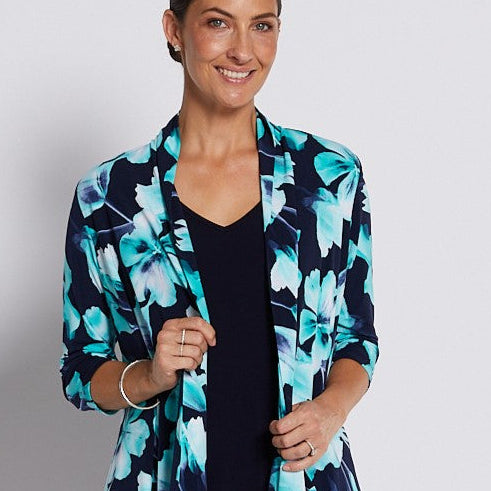 A woman modeling a navy blue cropped pants set with a navy and turquoise floral print blazer, a black top, and silver heeled sandals against a light gray studio background.