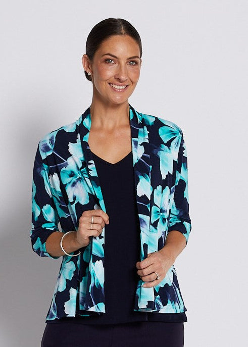 A woman modeling a navy blue cropped pants set with a navy and turquoise floral print blazer, a black top, and silver heeled sandals against a light gray studio background.