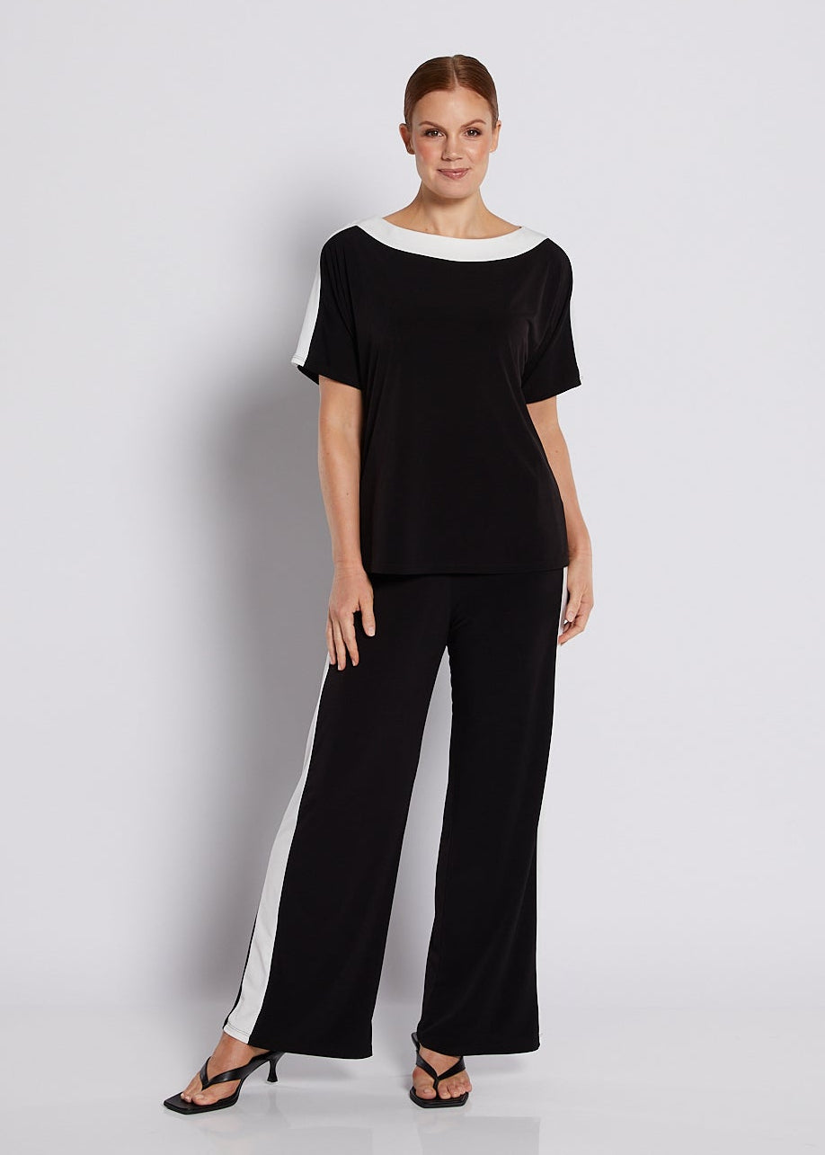 A woman modeling a black two-piece outfit consisting of a short-sleeve top with a white boat neckline and matching wide-leg pants with white side panels, standing against a white studio background.