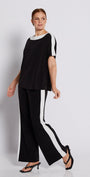 A woman modeling a black two-piece outfit consisting of a short-sleeve top with white side panels and white collar, paired with black wide-leg trousers featuring white side stripes, set against a minimalist white studio background.