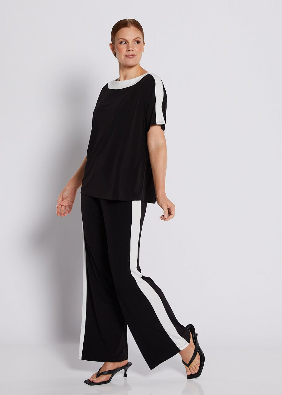 A woman modeling a black two-piece outfit consisting of a short-sleeve top with white side panels and white collar, paired with black wide-leg trousers featuring white side stripes, set against a minimalist white studio background.