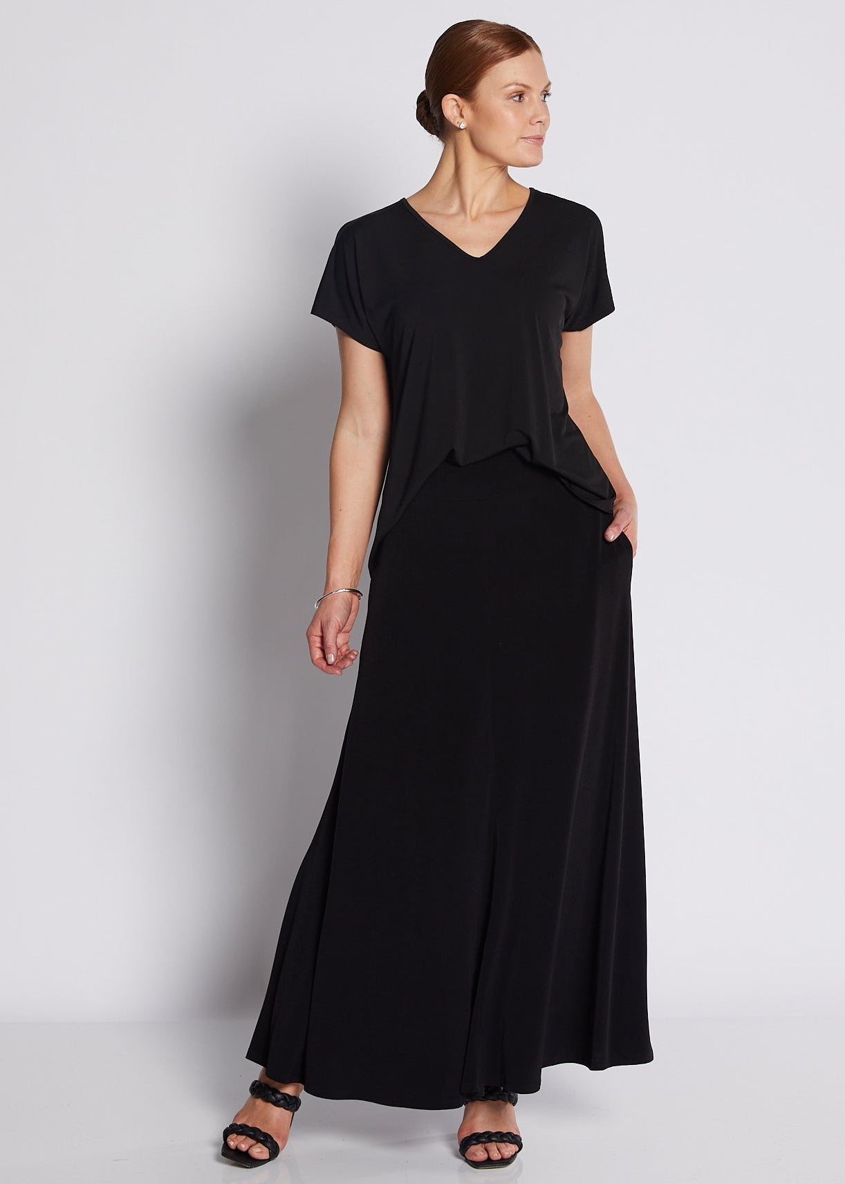 A woman modeling a black short-sleeve maxi dress with a V-neckline and side pockets, standing against a light gray studio background.