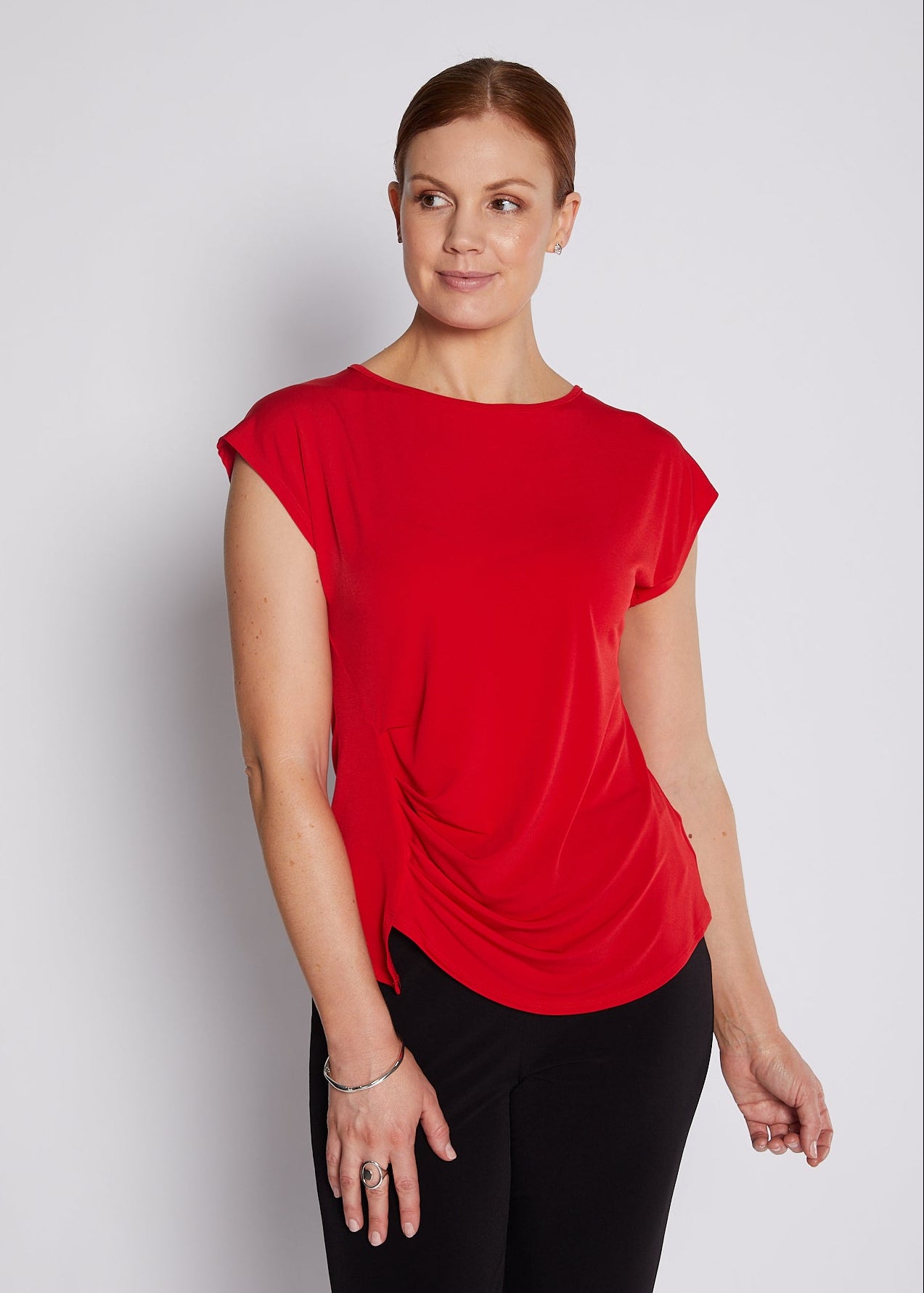 A woman modeling a bright red short-sleeve top with a draped, gathered front detail, paired with black pants, against a light gray studio background.