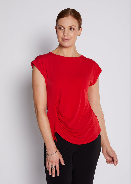 A woman modeling a bright red short-sleeve top with a draped, gathered front detail, paired with black pants, against a light gray studio background.