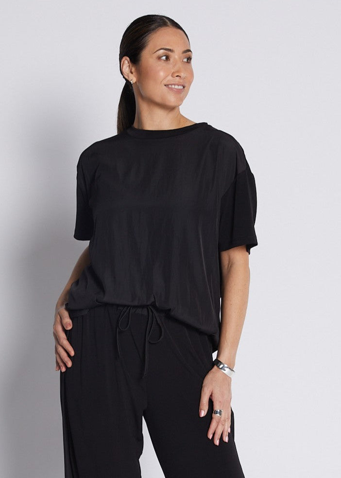 A woman modeling a black short-sleeve top and matching black wide-leg drawstring pants, paired with black slide sandals, against a light gray studio background.