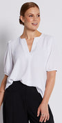 A woman modeling a white short-sleeve blouse with a split neckline and black wide-leg trousers, standing against a light gray studio background.