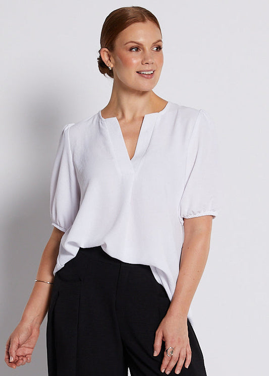 A woman modeling a white short-sleeve blouse with a split neckline and black wide-leg trousers, standing against a light gray studio background.