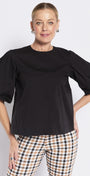 A woman modeling a black short puff-sleeve top with a crew neck, paired with beige and black checkered trousers, against a light gray studio background.