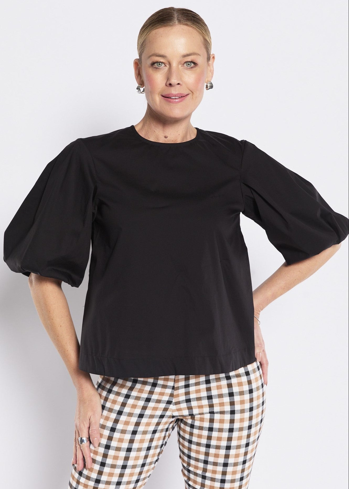A woman modeling a black short puff-sleeve top with a crew neck, paired with beige and black checkered trousers, against a light gray studio background.